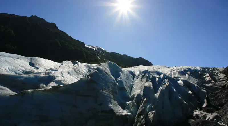 Sunlit ridges of blue-white ice on the surface of Exit Glacier below a dark mountainside in Kenai Fjords National Park