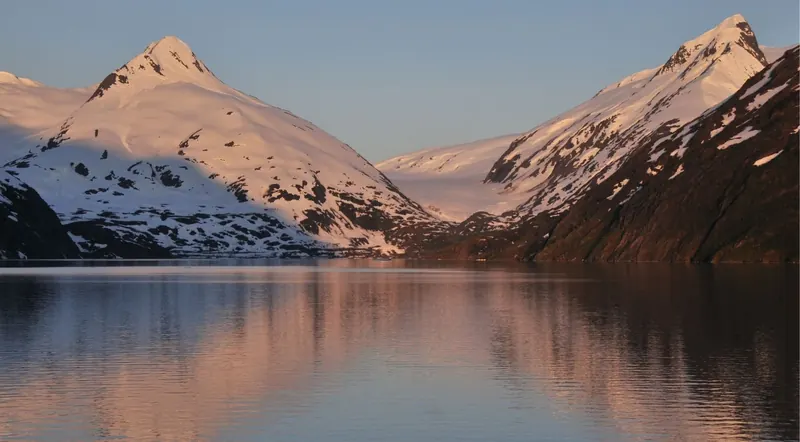 Pink alpenglow on snow-covered peaks reflected in the still water of an Alaskan fjord at sunset near Seward